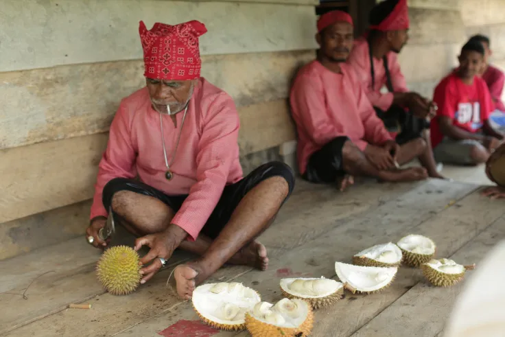 Pria berikat kepala merah membuka durian di lantai kayu bersama warga